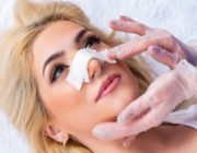 Young woman lies down while Plastic Surgeon touches nose bandages to assess Rhinoplasty results