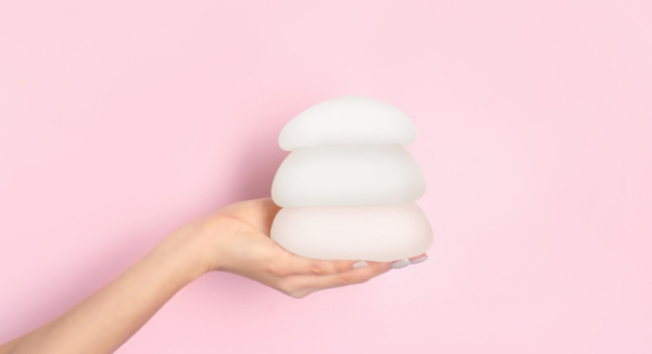 woman holds 3 sizes of breast implants against pink background.